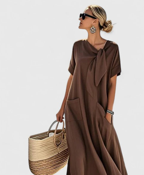 Woman wearing a flowing brown Summer Linen Dress with asymmetrical neck tie detail and side pockets, styled with sunglasses and woven basket bag